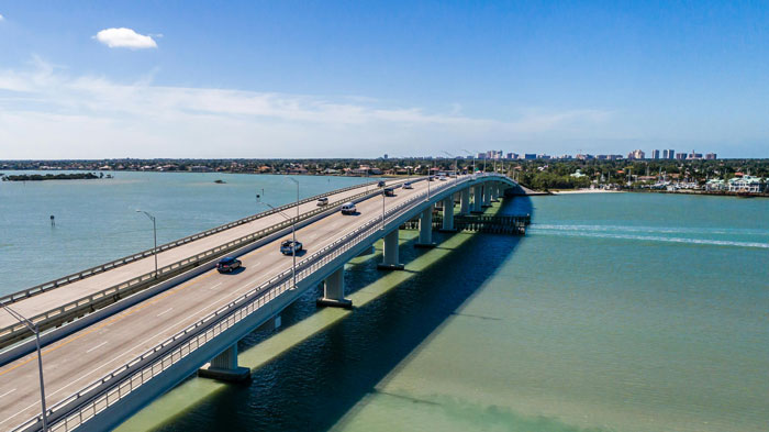 rial view of a bridge over turquoise waters connecting the mainland to a coastal island with a clear blue sky overhead.