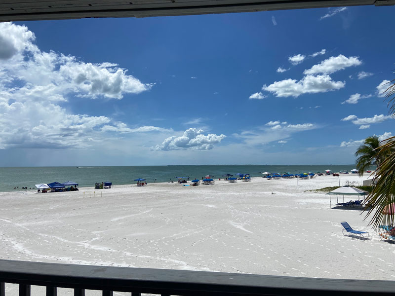 View from a balcony overlooking a white sandy beach with beachgoers and blue parasols, and a vast ocean extending to the horizon