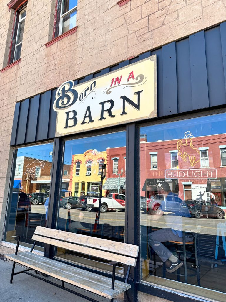 Vintage 'Born in a Barn' sign above a bench on a storefront, reflecting the street and other buildings in the window.