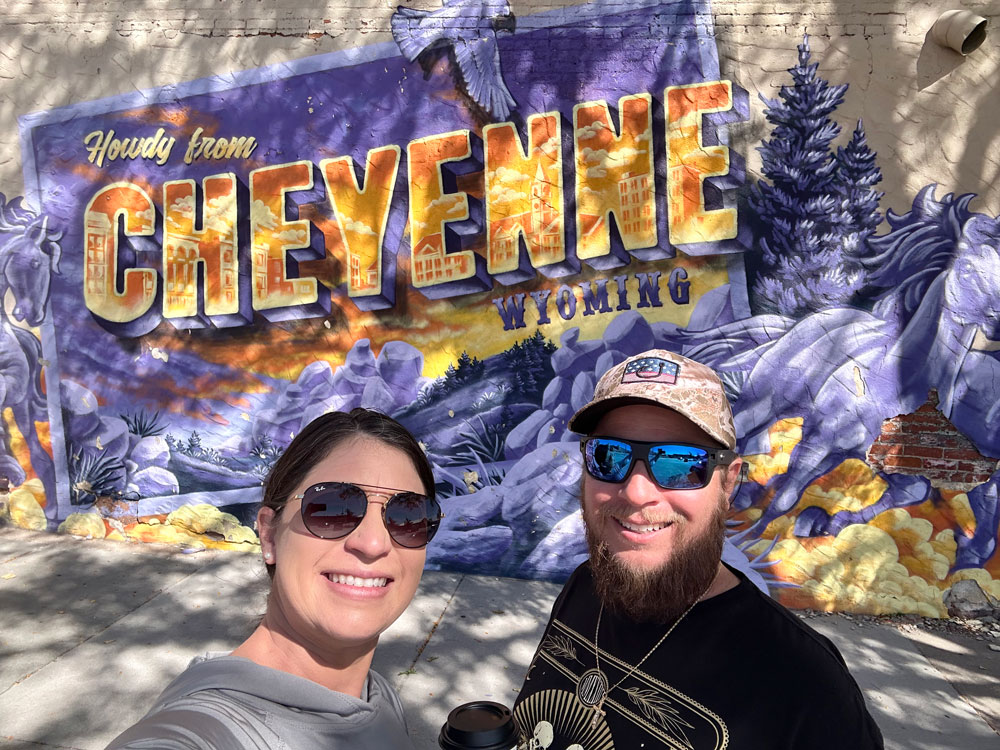 A couple taking a selfie in front of a vibrant street mural that reads 'Howdy from CHEYENNE WYOMING'