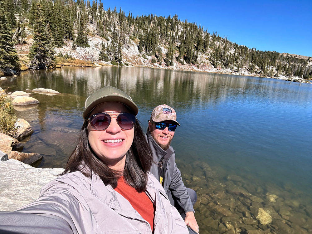 A smiling couple takes a selfie with a crystal clear mountain lake and forested slopes in the background