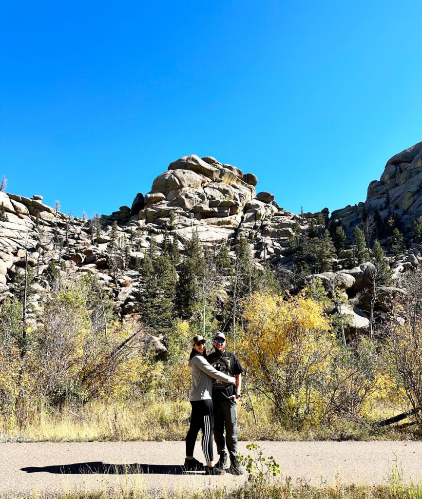 A couple posing for a photo at Vedauwoo with unique rock formations surrounded by autumn-colored foliage under a clear sky