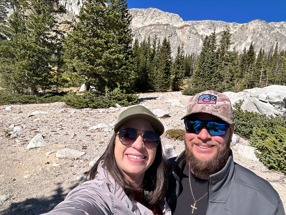 Smiling couple taking a selfie with the rugged wilderness of Medicine Bow National Forest featuring pine trees and rocky mountain peaks in the background.