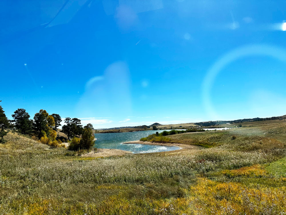 Scenic view of Curt Gowdy State Park with a small lake surrounded by grassy fields and trees under a bright blue sky with lens flare.