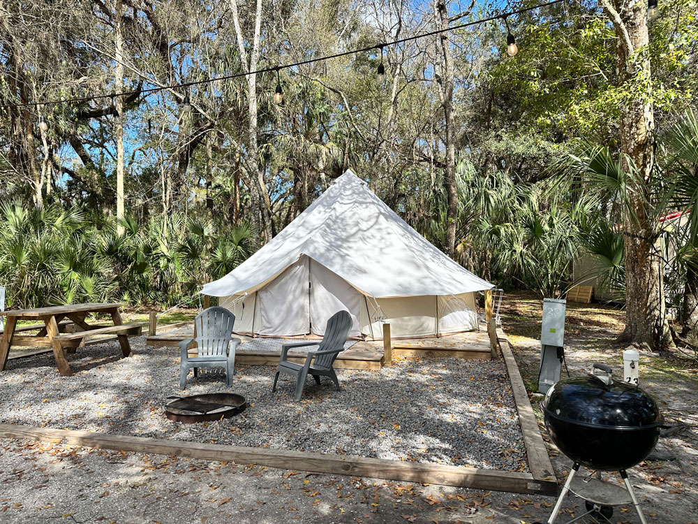 A glamping site with a large white tent, wooden picnic table, and two plastic chairs arranged on a gravel surface, surrounded by lush trees and a portable barbecue grill.
