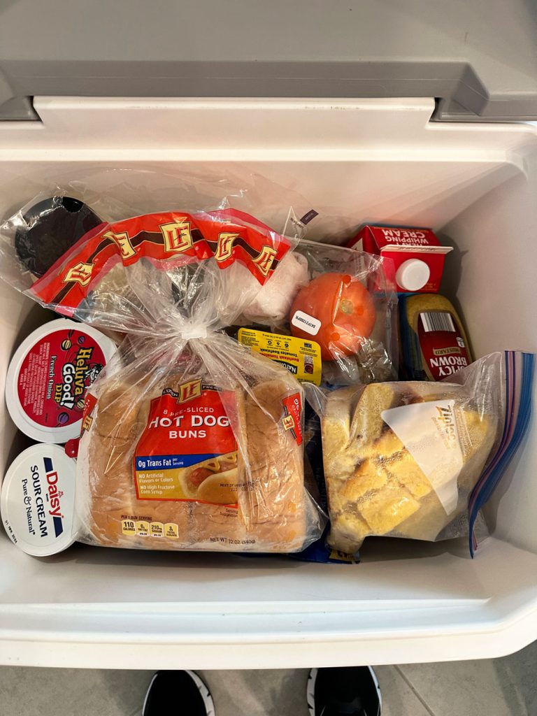 Open cooler packed with an assortment of groceries, including hot dog buns, sour cream, a tomato, mustard, and a bag with cornbread