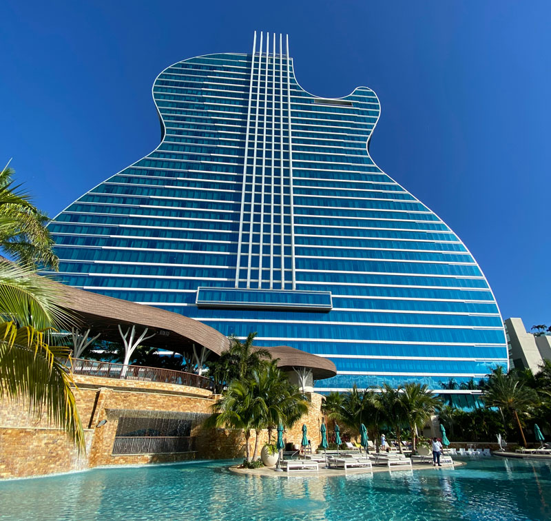 The iconic guitar-shaped hotel at the Hard Rock in Florida with a sprawling pool area and tropical palm trees under a clear blue sky.