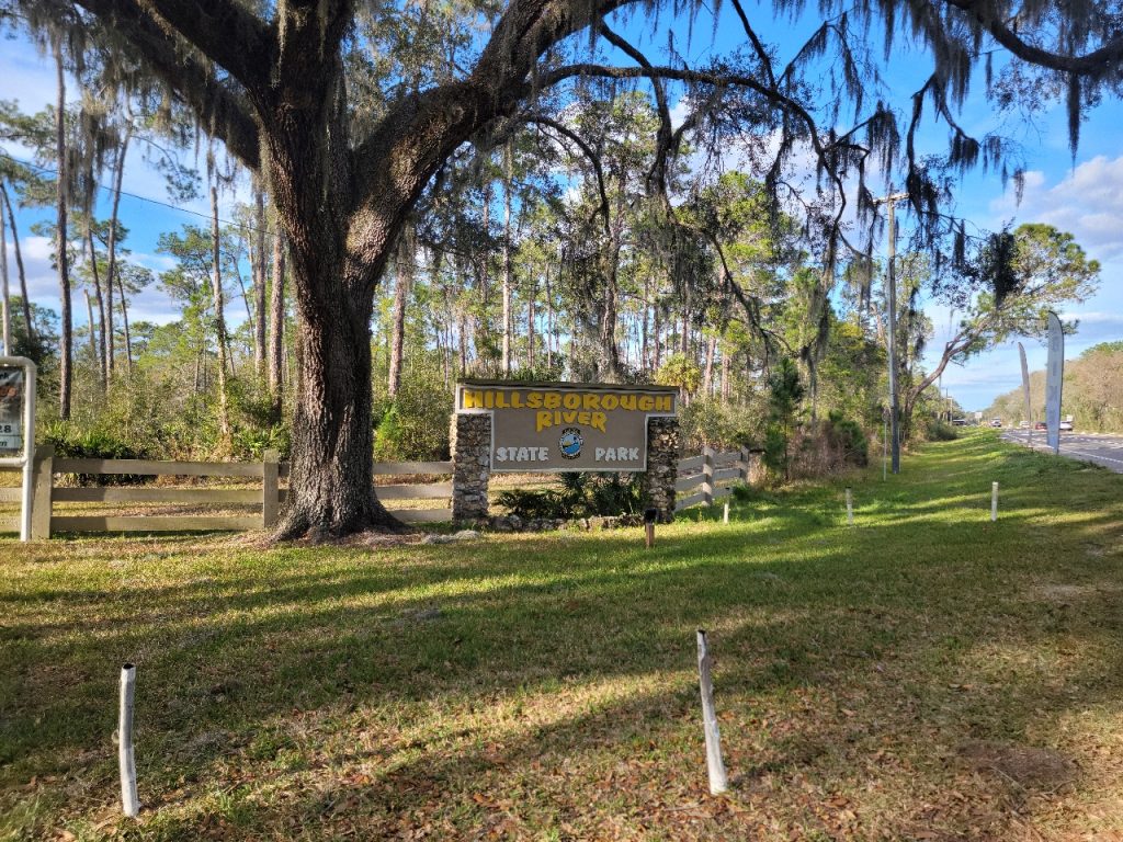 entrance sign of 'Hillsborough River State Park' with a large tree and Spanish moss in the foreground, clear blue sky above.