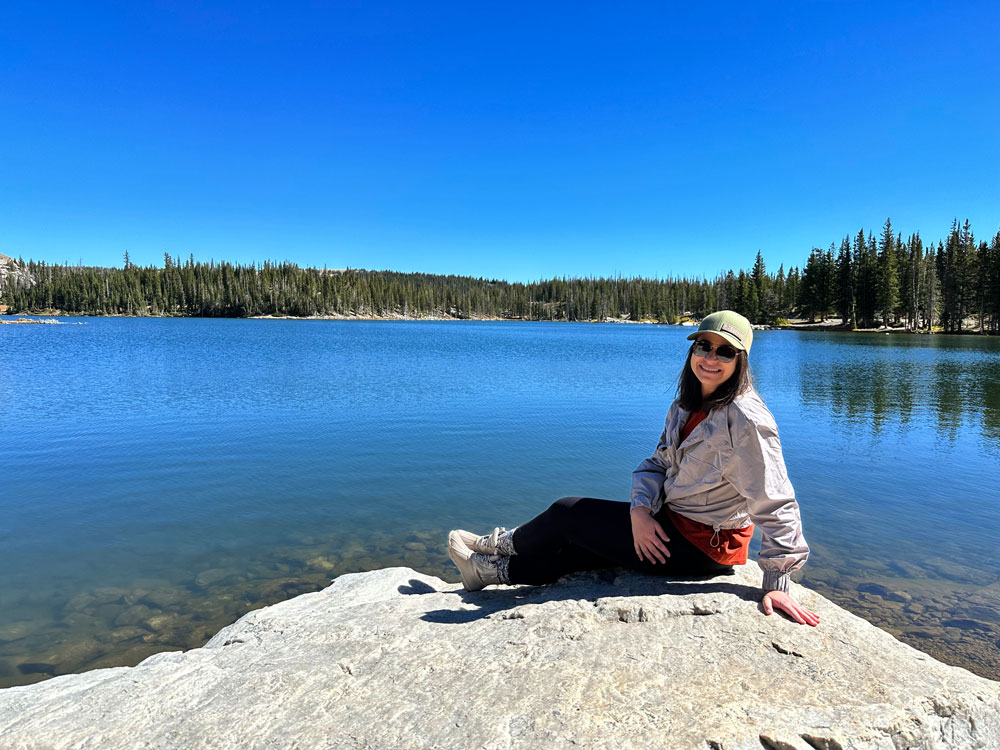 A woman in a cap and sunglasses sitting on a rock by a serene mountain lake surrounded by pine trees under a clear blue sky