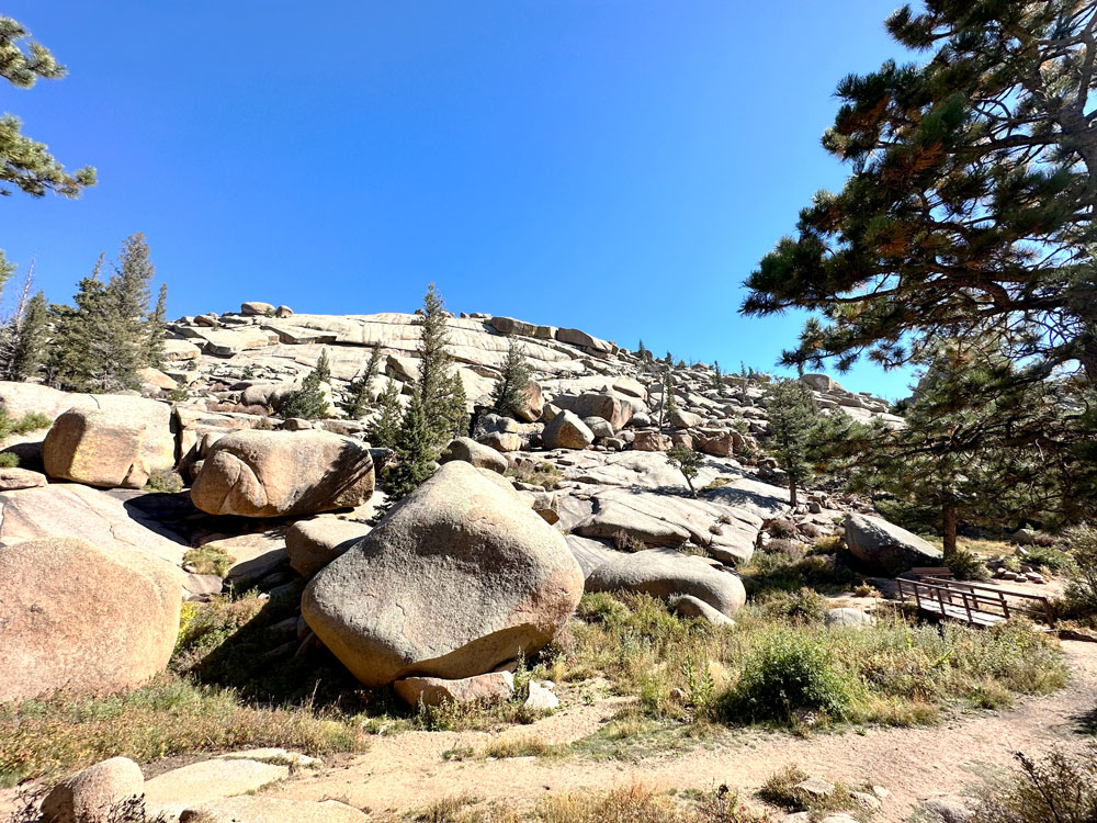 Rocky landscape featuring large boulders, pine trees, and a wooden walkway under a bright blue sky in Vedauwoo Wyoming