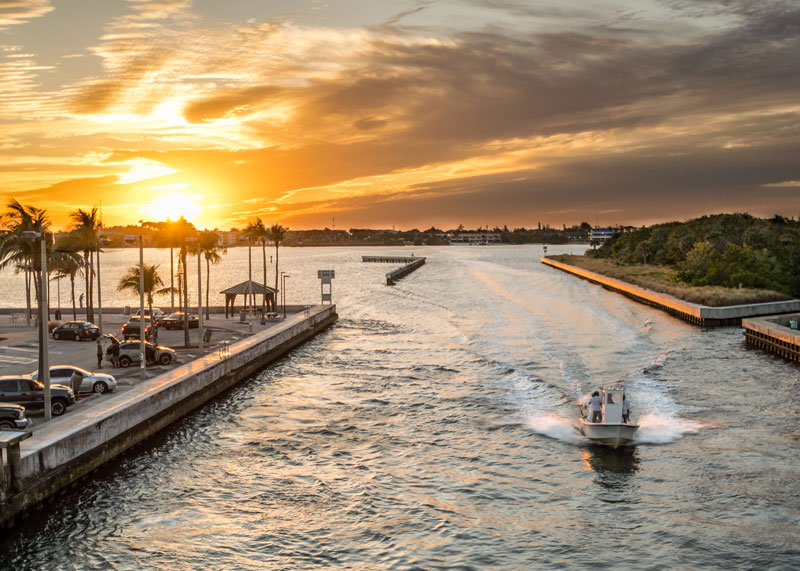 A speedboat cruises through a water channel at sunset, with the golden sun dipping below the horizon and silhouetting palm trees along the waterfront