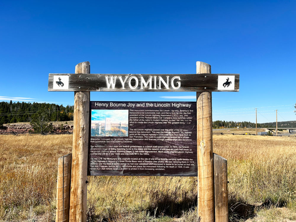 Wooden signpost with 'WYOMING' and a detailed informational board about Henry Bourne Joy and the Lincoln Highway against a natural landscape