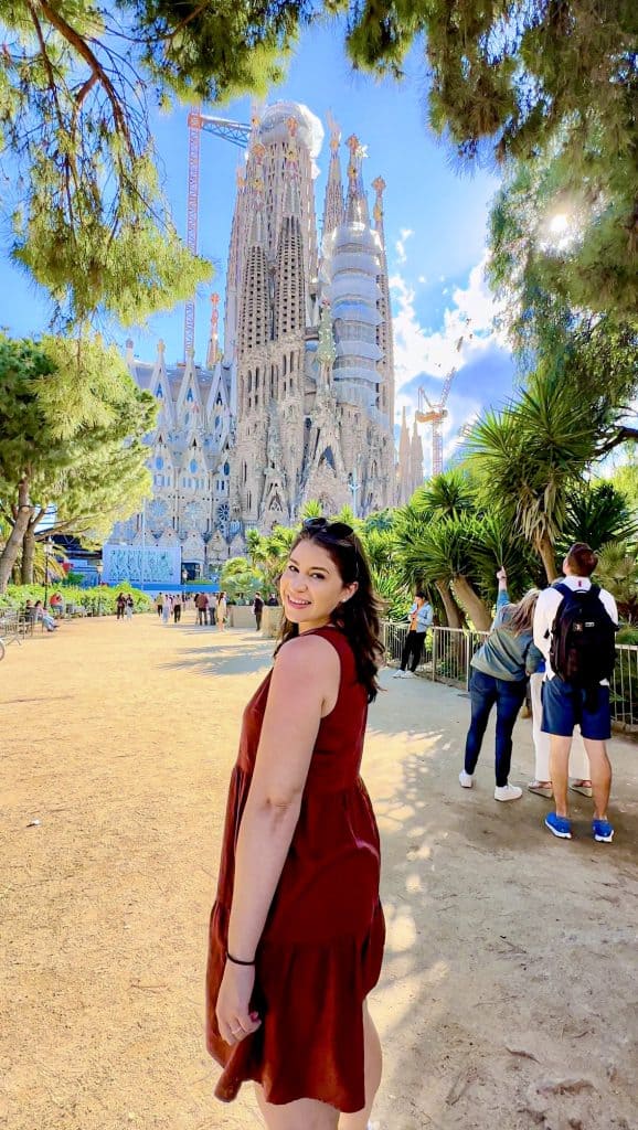A woman in a maroon dress smiles in front of the Sagrada Família in Barcelona, surrounded by greenery under a blue sky.