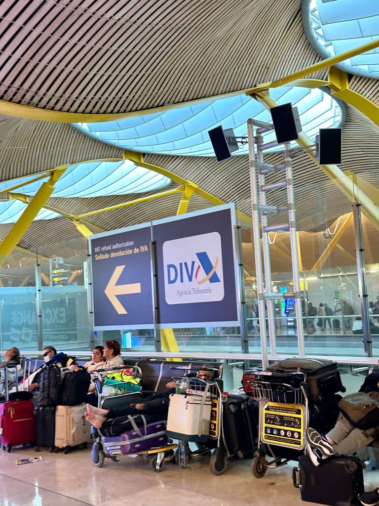  Travelers with luggage resting near a large sign for "DIVA" VAT Refund Authorization in an airport. The curved architectural ceiling with large yellow supports is prominently featured.