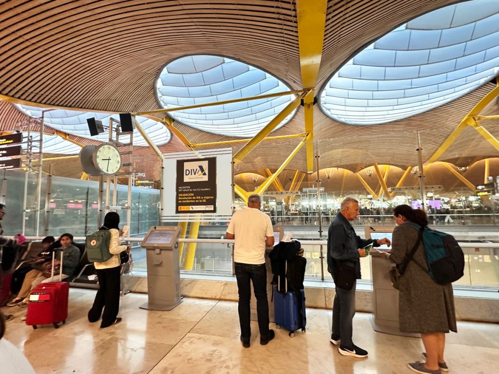 Passengers at an airport using a VAT refund kiosk under a large curved ceiling with yellow beams. A clock and seating area are visible in the background.