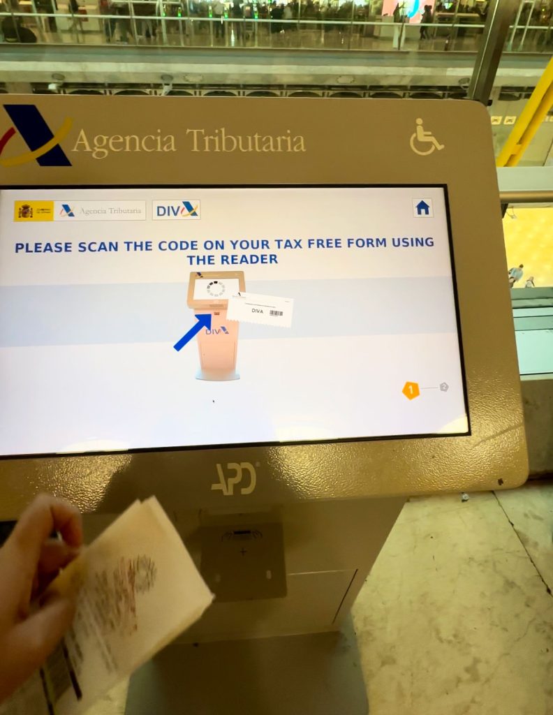 A person’s hand holding a tax-free form near a VAT refund kiosk screen, ready to scan the barcode. The screen displays instructions for scanning.