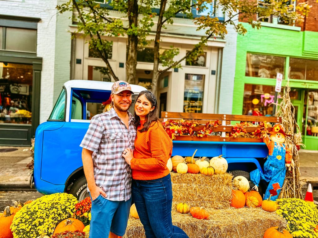 A smiling couple posing by a bright blue truck decorated with pumpkins and autumn leaves on hay bales, set on a quaint town street.