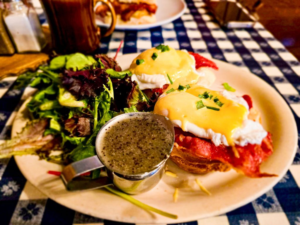 Plate featuring eggs Benedict with a side of leafy greens and a small cup of creamy dressing, set on a checkered tablecloth.