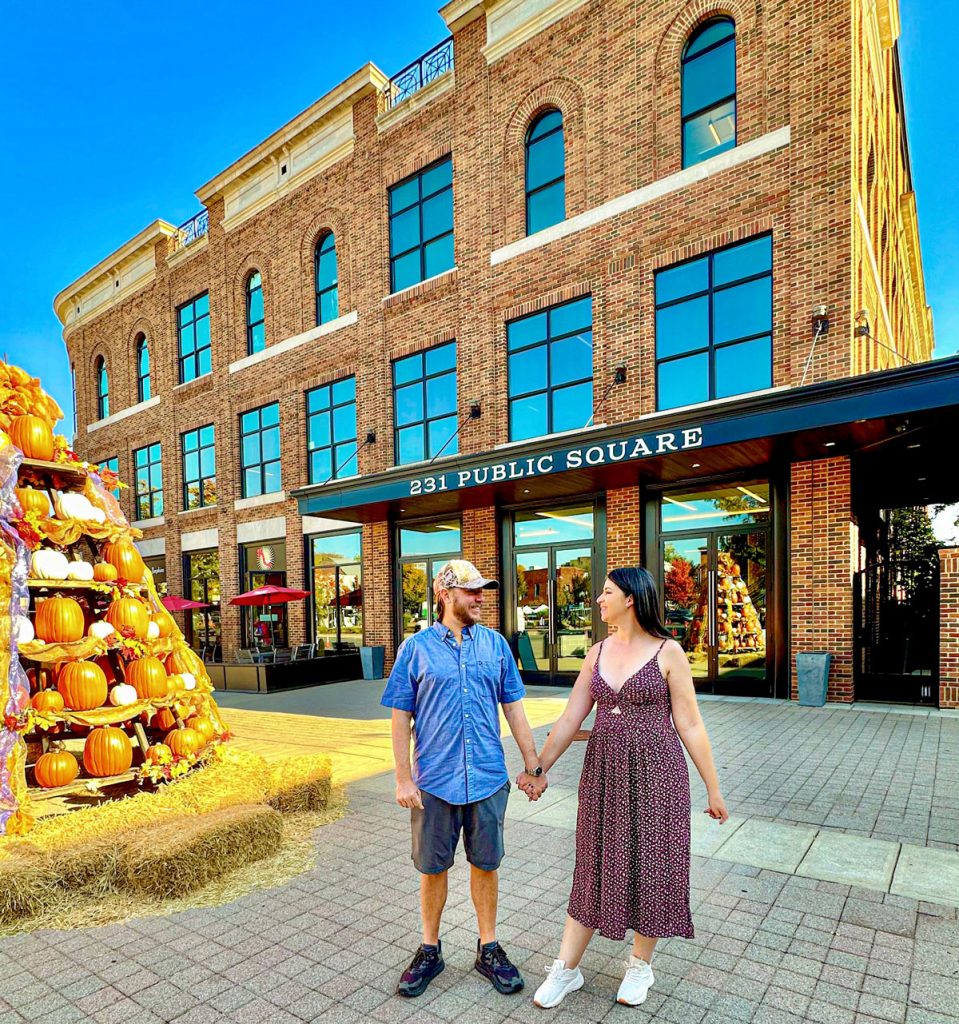 A couple holding hands and smiling at each other in front of a building labeled "231 Public Square," next to a large decorative display of pumpkins.
