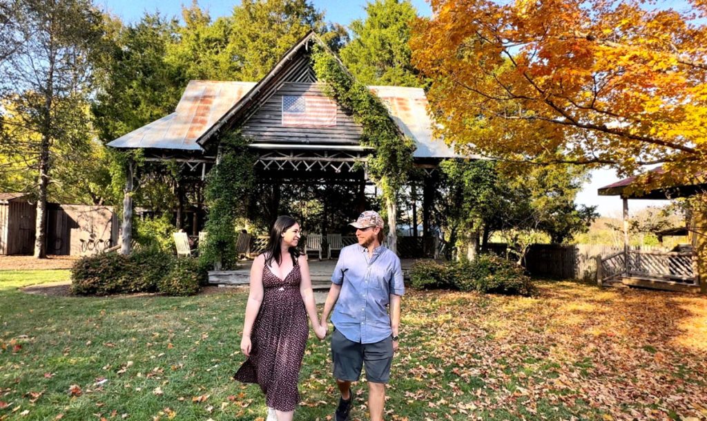 A couple holding hands and smiling while walking through a garden with an ivy-covered pavilion in the background.
