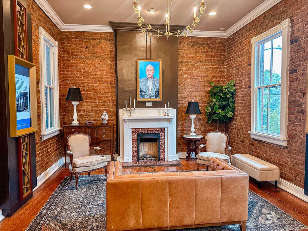 A cozy, vintage-style lounge room with exposed brick walls, classic furniture, a chandelier, and a fireplace with a portrait above it.