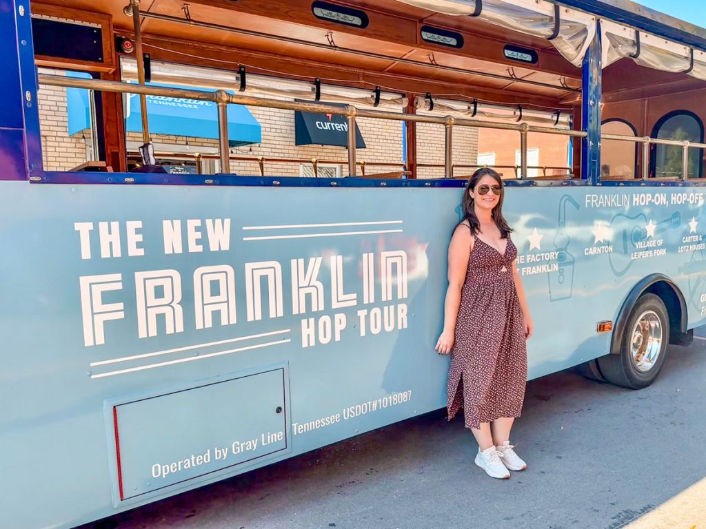 Woman standing in front of a blue "Franklin Hop-On, Hop-Off" tour bus, smiling on a sunny day.