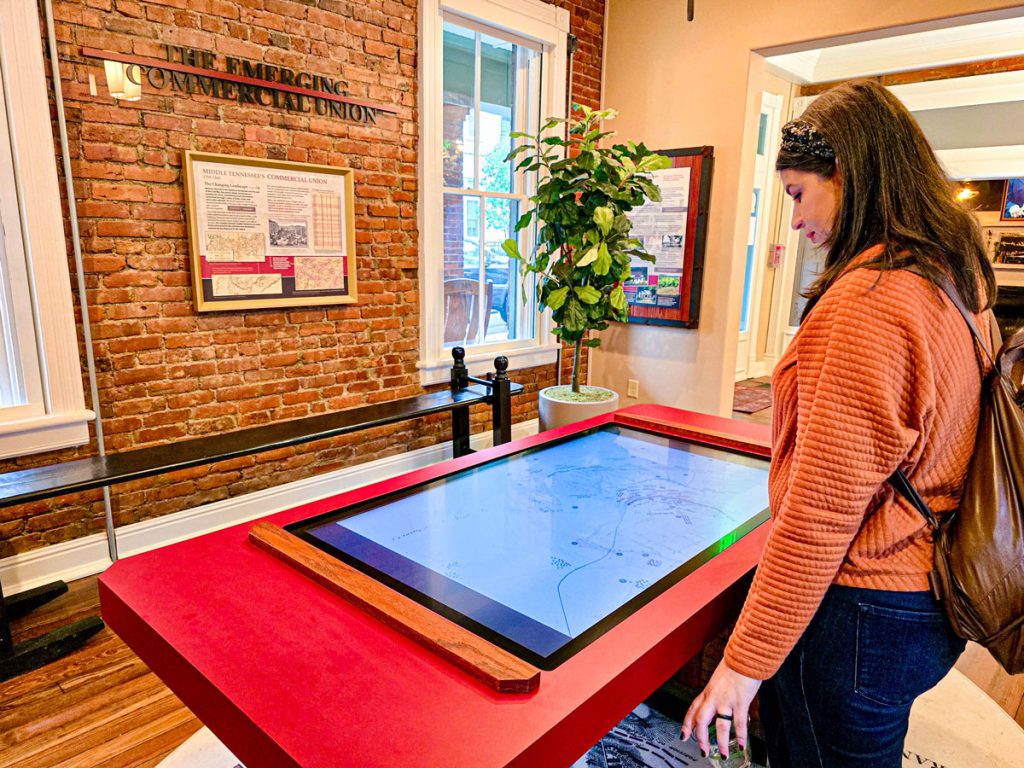 A visitor interacts with a large touchscreen display in a museum exhibit, surrounded by exposed brick walls and historical displays.