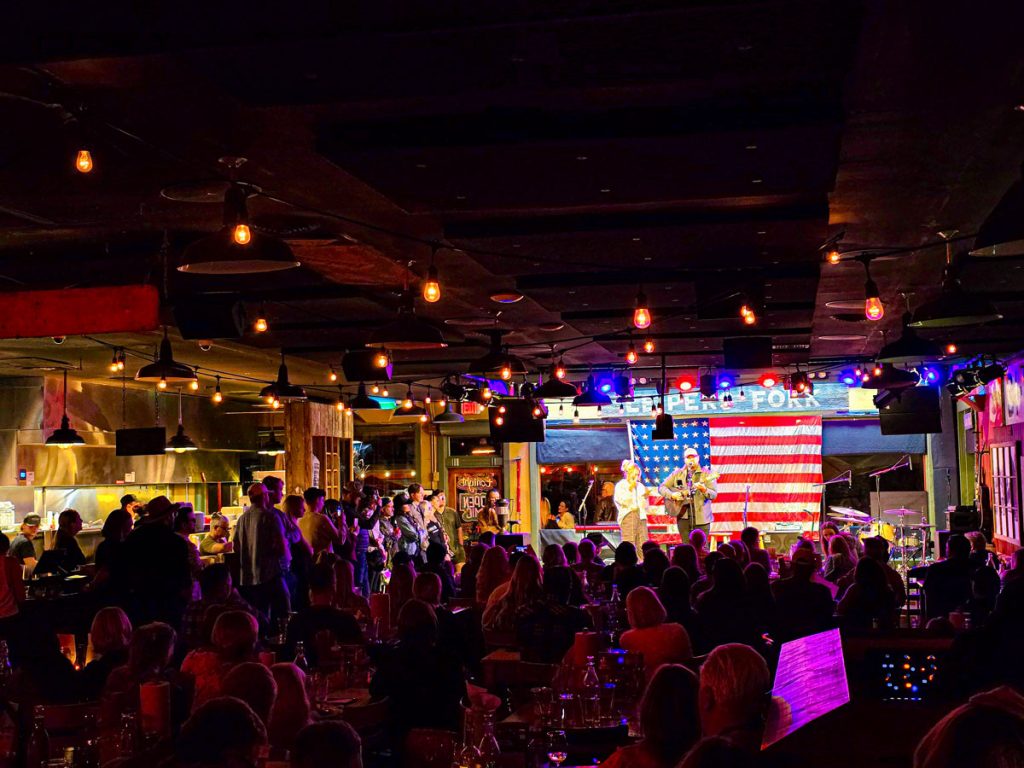 A vibrant live music performance on a stage adorned with an American flag, viewed by an audience in a warmly lit venue.
