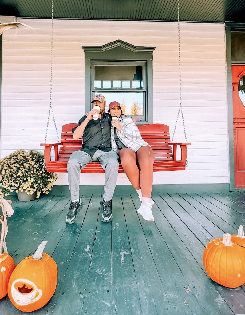 A couple sitting on a red porch swing, sipping from cups and surrounded by pumpkins, enjoying a cozy autumn afternoon.