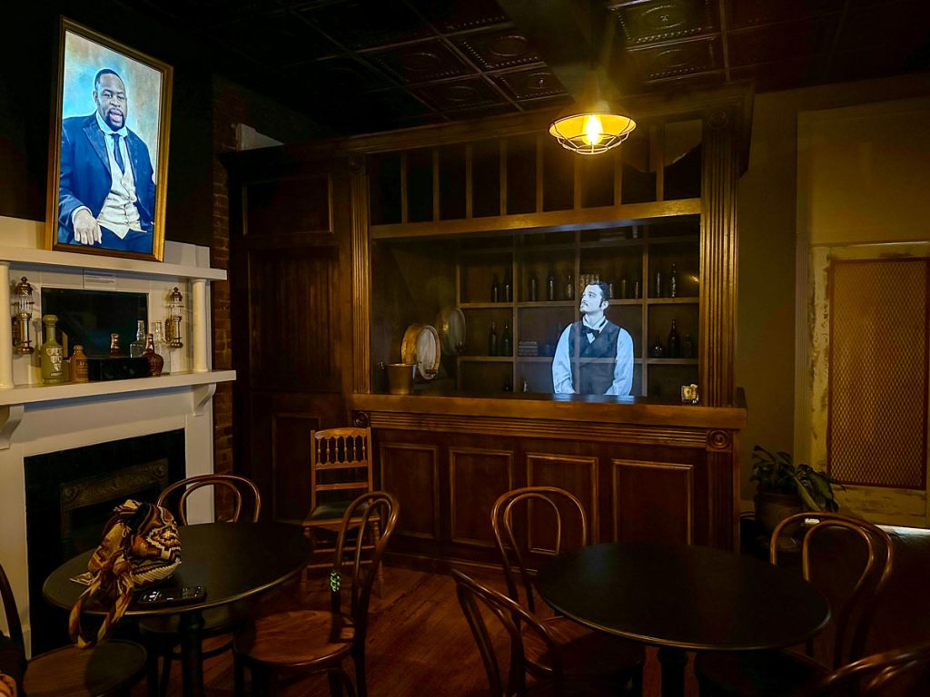 Dimly lit, historic-style room featuring a wooden bar with a holographic bartender and a portrait above a white mantel.